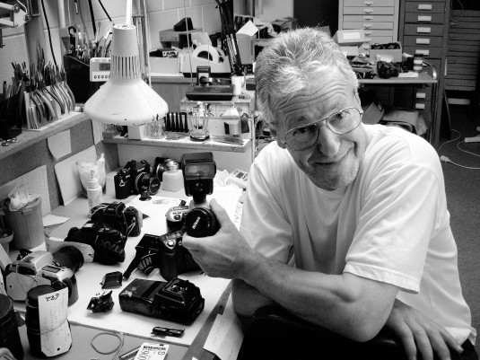 Black & white photo of Phil Jacobs at his workshop desk, which is covered in cameras in various states of disassembly.