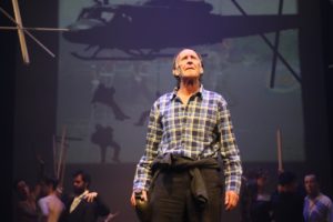 An older man stands centre stage. Behind him is a backdrop of soldiers rappelling from a helicopter.