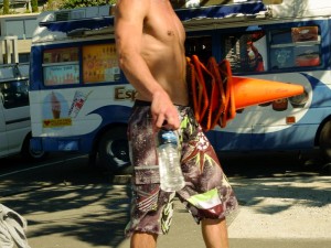 A headless shot of a topless man in boardshorts holding bottled water and 6 traffic cones in Oriental Bay.