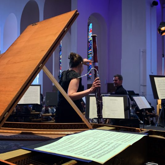 Behind a harpsichord, a bassoonist talks to a colleague, with Händel scores on music stands.