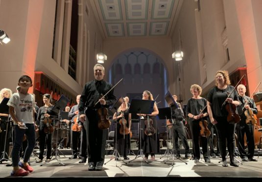 Vesa-Matti Leppänen and the orchestra prepare to take a bow, while a round boy holding a toy airliner has climbed the stage in appreciation.