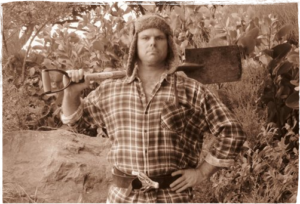 Sepia coloured photo of Geenty in a checkshirt, hat with ear flaps and a spade across his shoulders