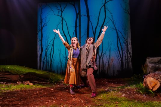 A photo of a man and woman, standing, smiling and gesturing wildly, in front of a woods backdrop.