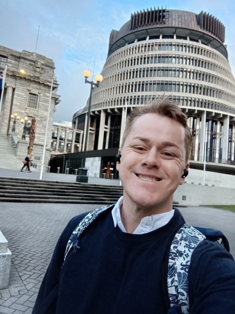 Joel, a Pākeha man, stands outside the beehive and smiles