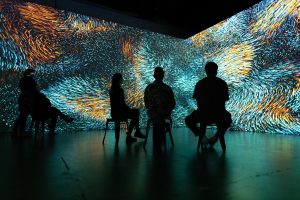 Silhouettes of three people on chairs in front of large projected image of digital animation called Rivers of Wind 