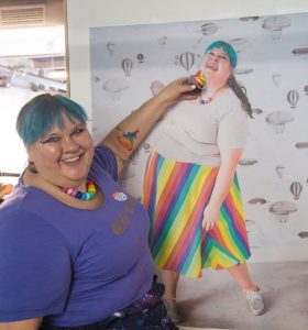 Fat babe Joanna tries to feed a rainbow cupcake to a large poster of herself in a rainbow skirt. 