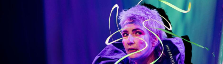 A pākehā woman's face wearing clown makeup. A neon ribbon cascades in front of their face.