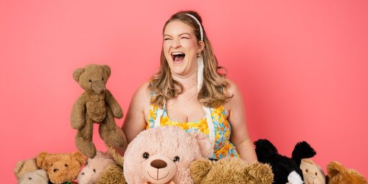 Blonde woman wearing a floral dress laughing while surrounded by teddy bears.