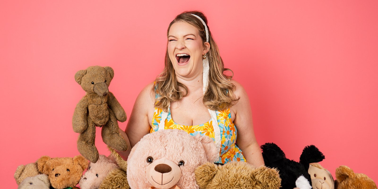 Blonde woman wearing a floral dress laughing while surrounded by teddy bears.