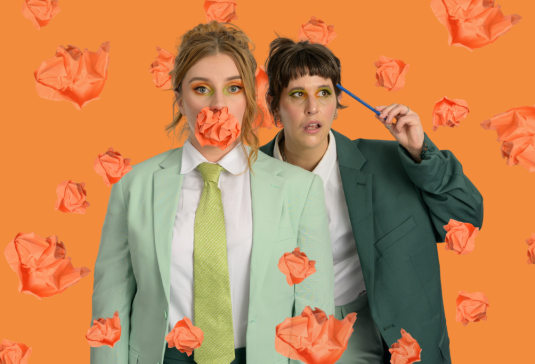 To adults stand in business suits. One has an orange intheir mouth, the other raising a pencil to their head.